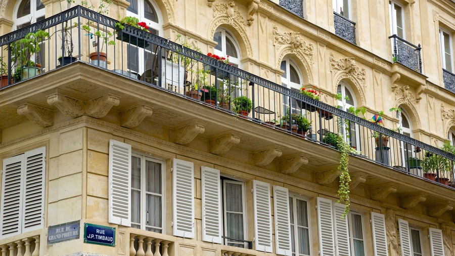 Un bâtiment à l’architecture richement décorée, doté de volets blancs et de balcons ornés de plantes en pot.