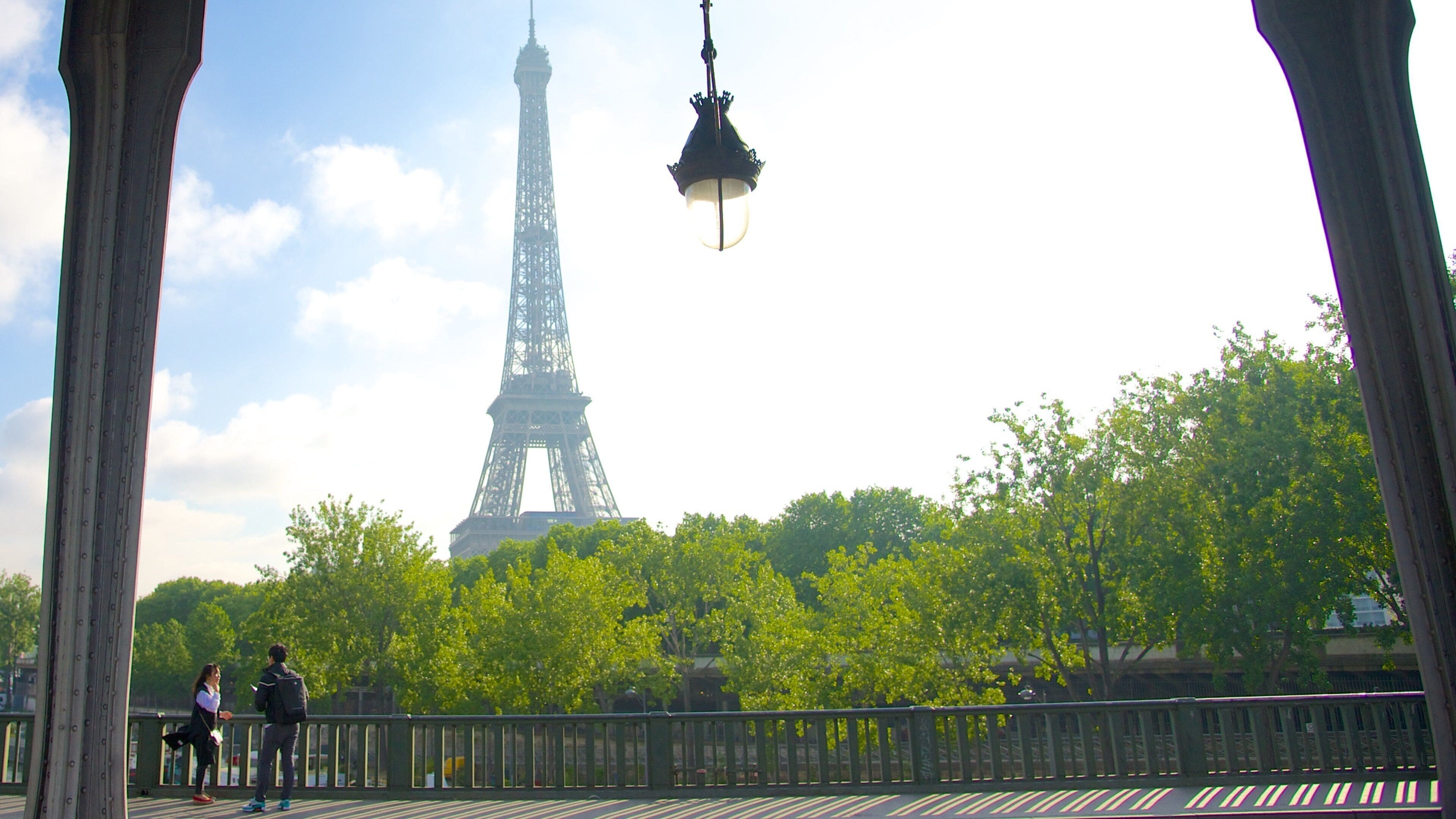 A Torre Eiffel com uma linha verde de árvores em primeiro plano e uma cerca de metal.