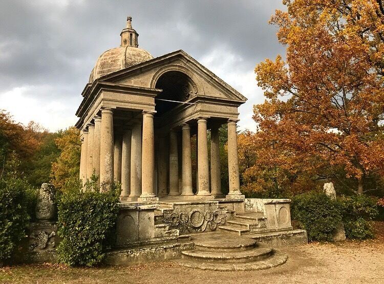 Ein klassizistisches Gebäude mit Säulen und einer Kuppel, umgeben von Herbstbäumen und einer Steinsitzbank.