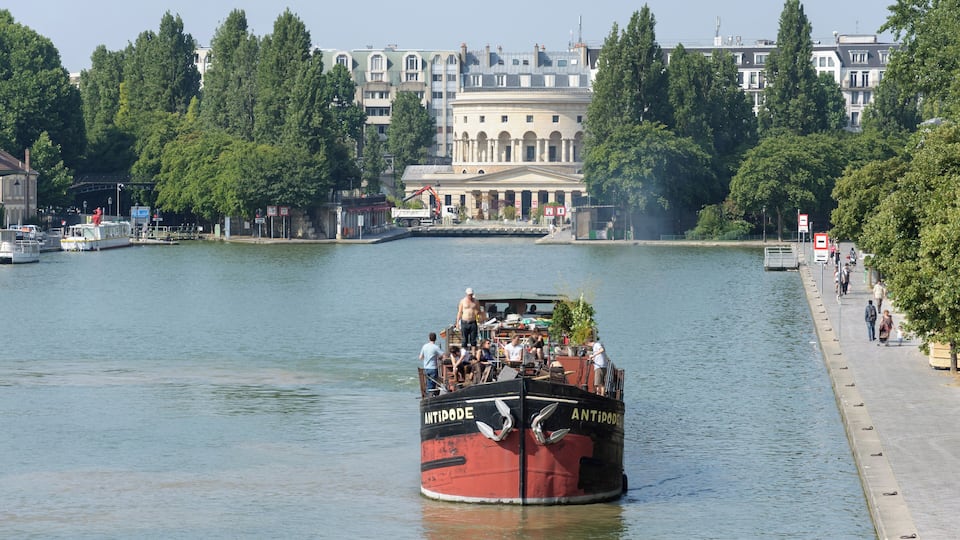 Um barco vermelho chamado "ANTIPODE" num rio calmo, rodeado por árvores e edifícios.