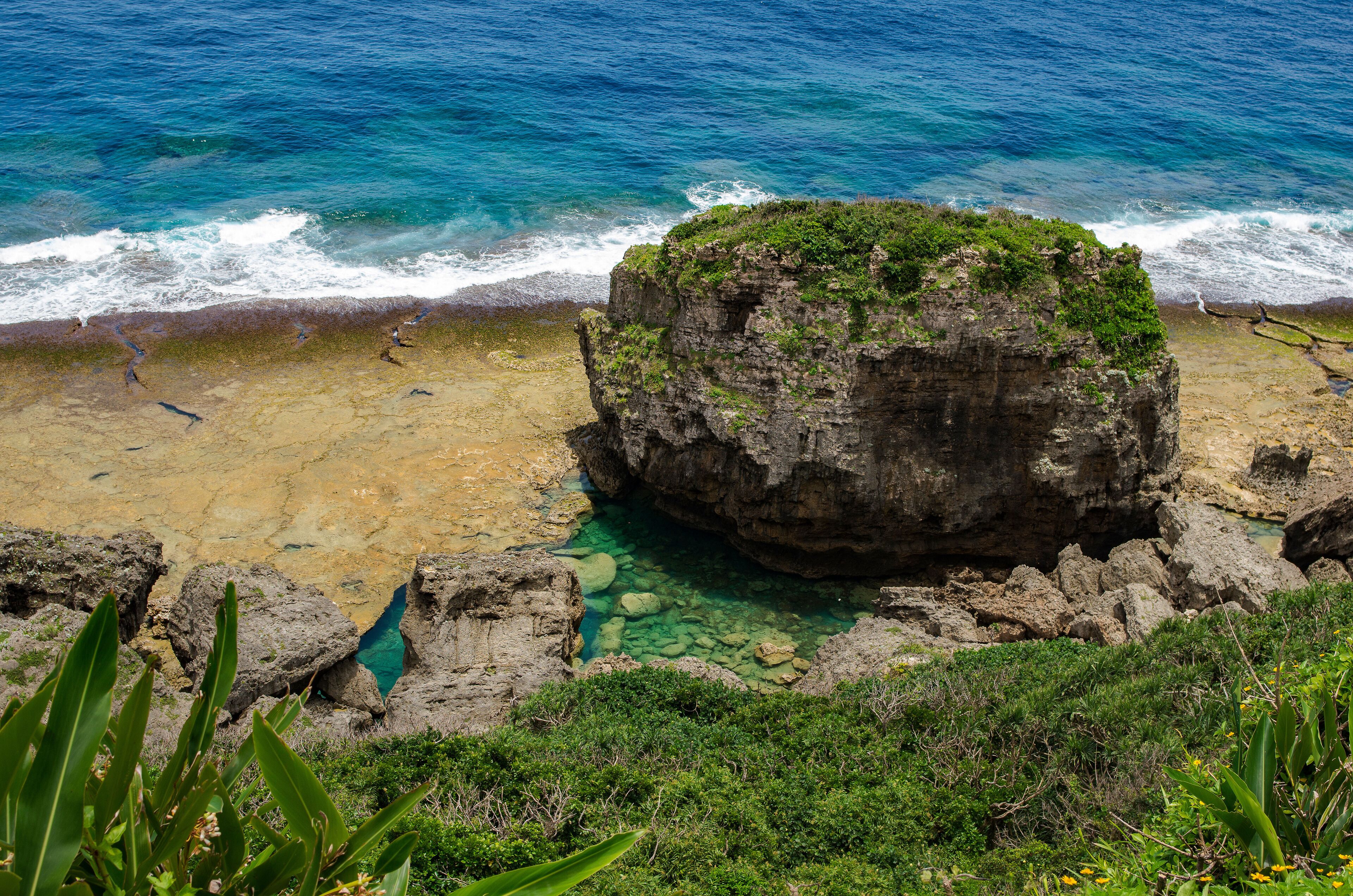 Una costa rocosa con aguas azul turquesa, una gran formación rocosa y vegetación verde.
