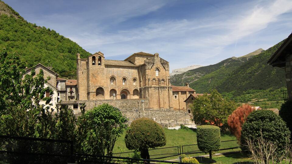 A historic stone building with arched windows and a bell tower, surrounded by lush green hills and trees.