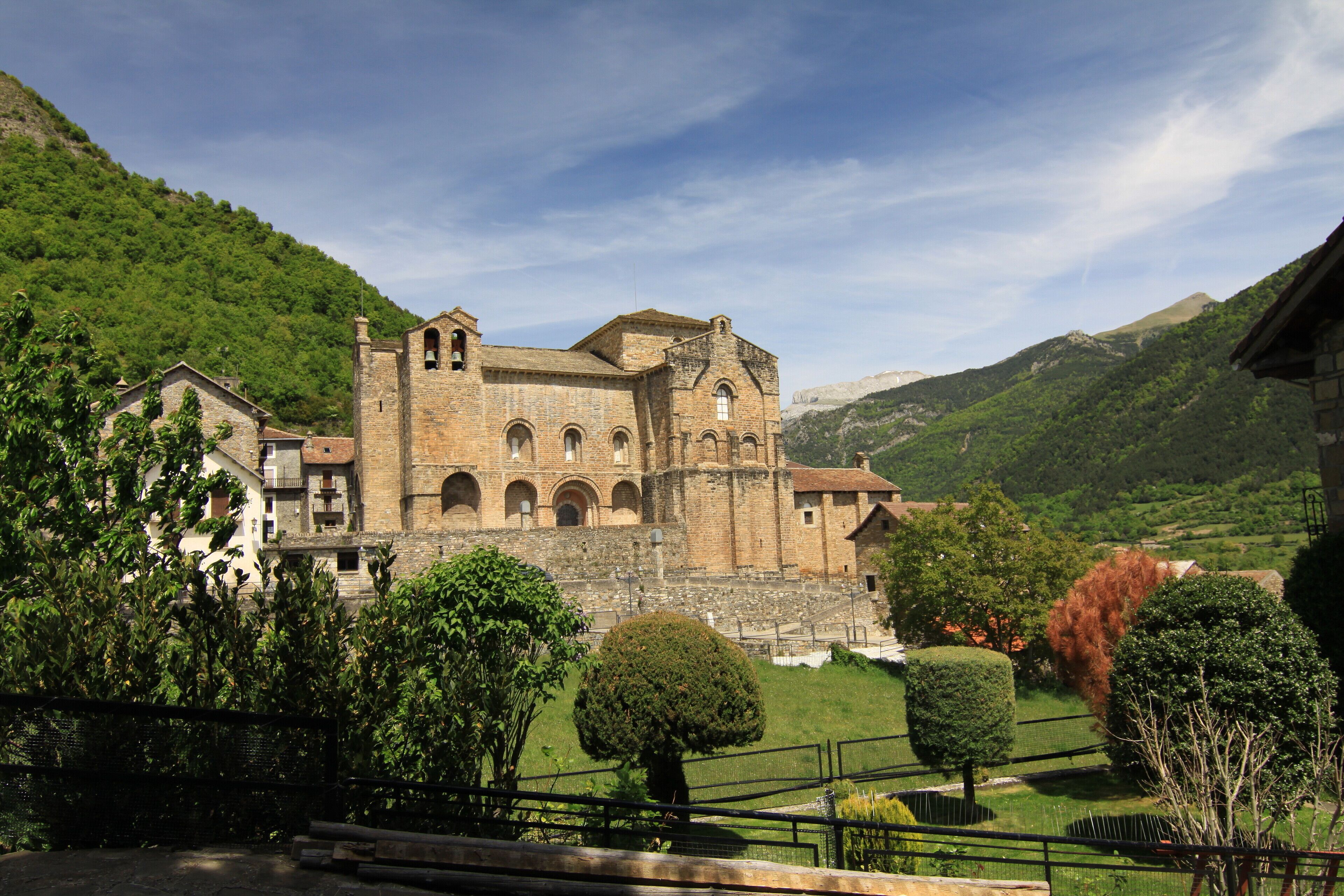 A historic stone building with arched windows and a bell tower, surrounded by lush green hills and trees.
