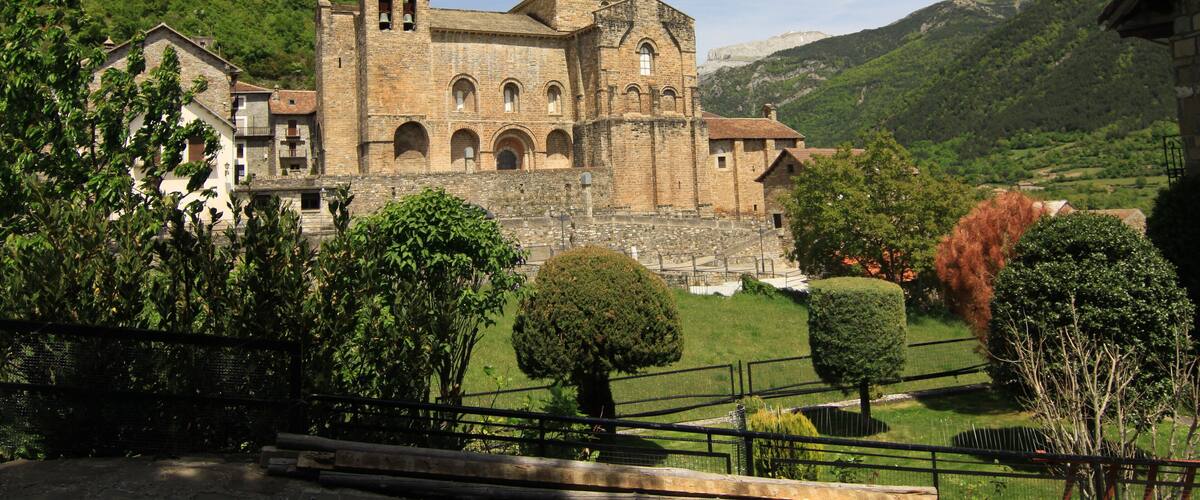 A historic stone building with arched windows and a bell tower, surrounded by lush green hills and trees.