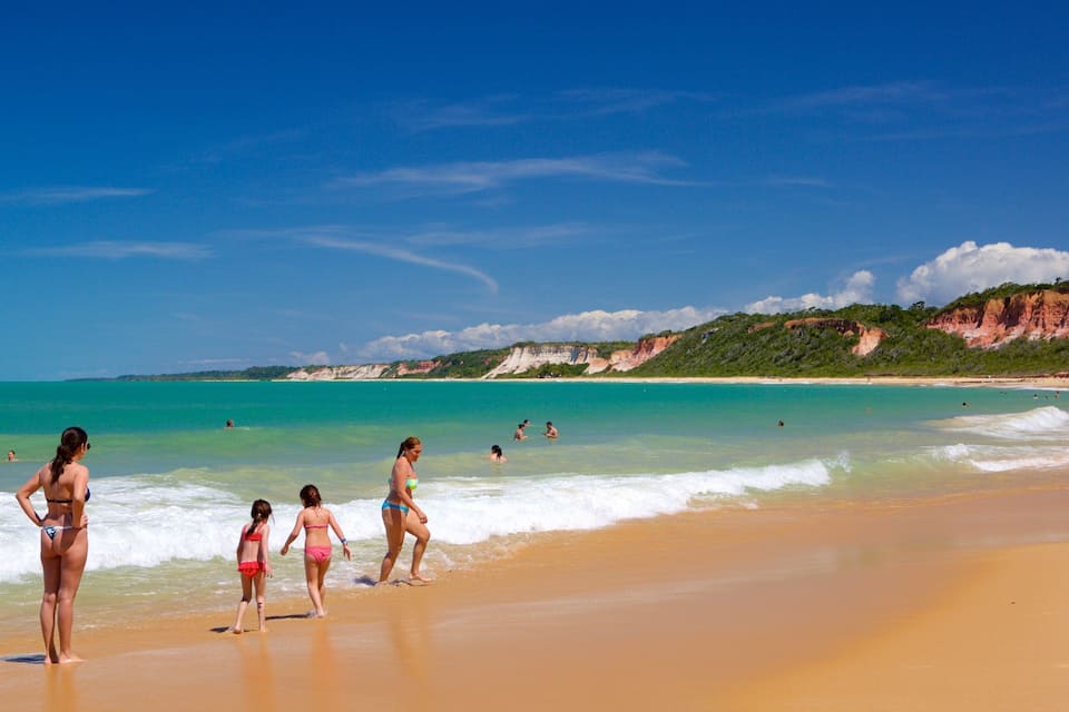Una playa con agua turquesa cristalina, orilla arenosa y acantilados rocosos al fondo.