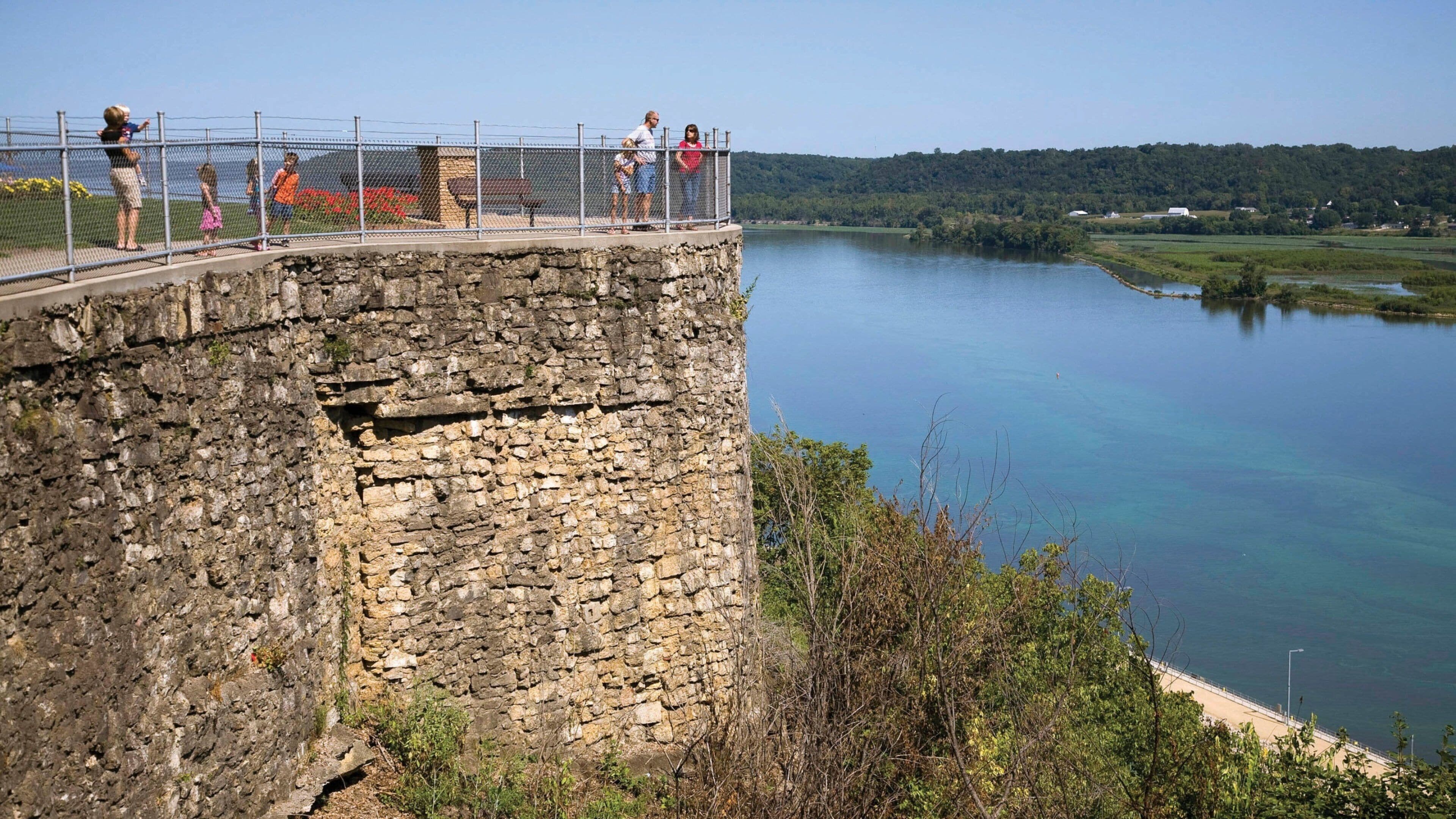 A stone bridge with people walking over it, overlooking a calm lake and a forested hill in the background.