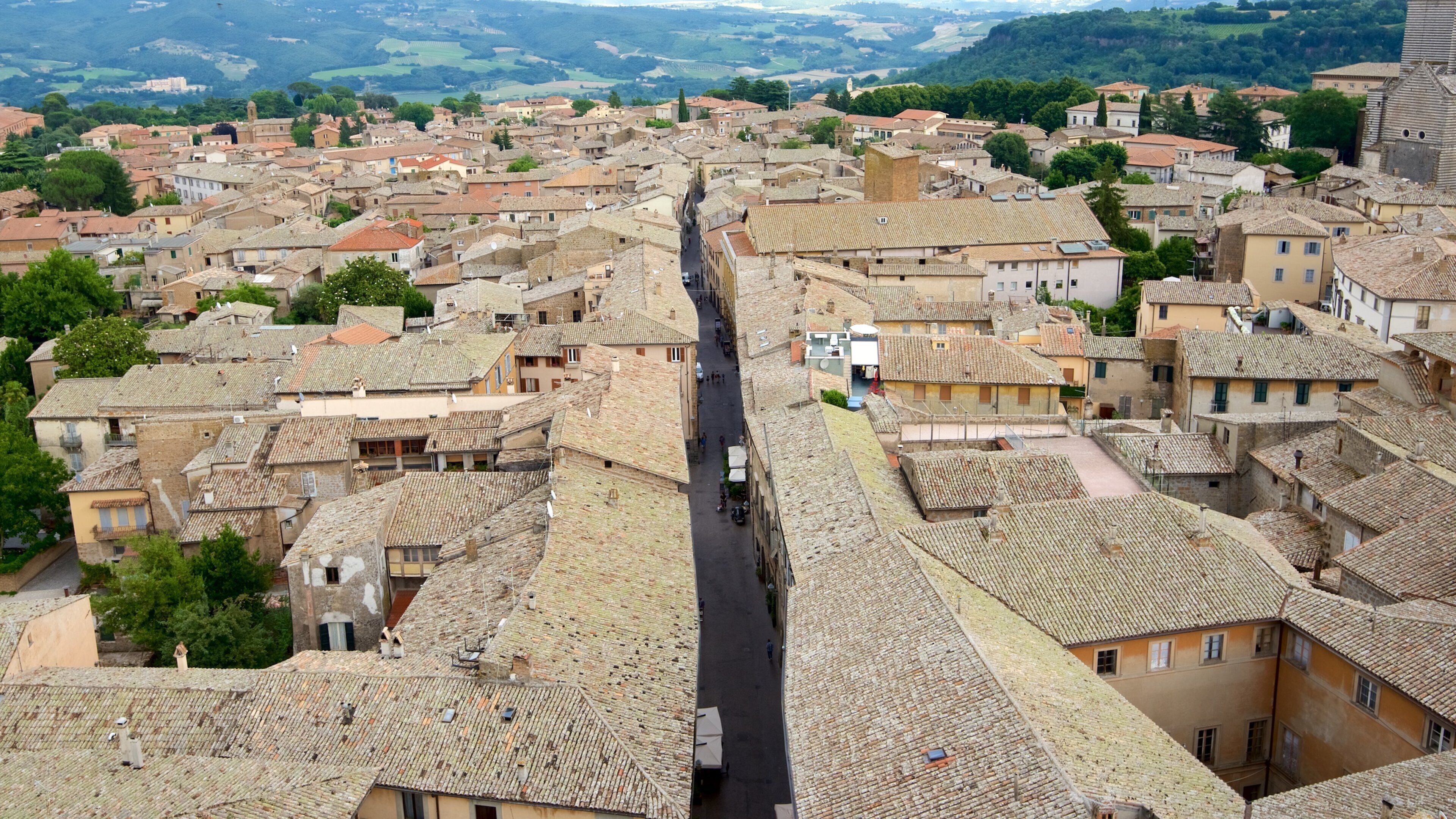 A historic town with terracotta-roofed buildings, a central street, and surrounding hills.
