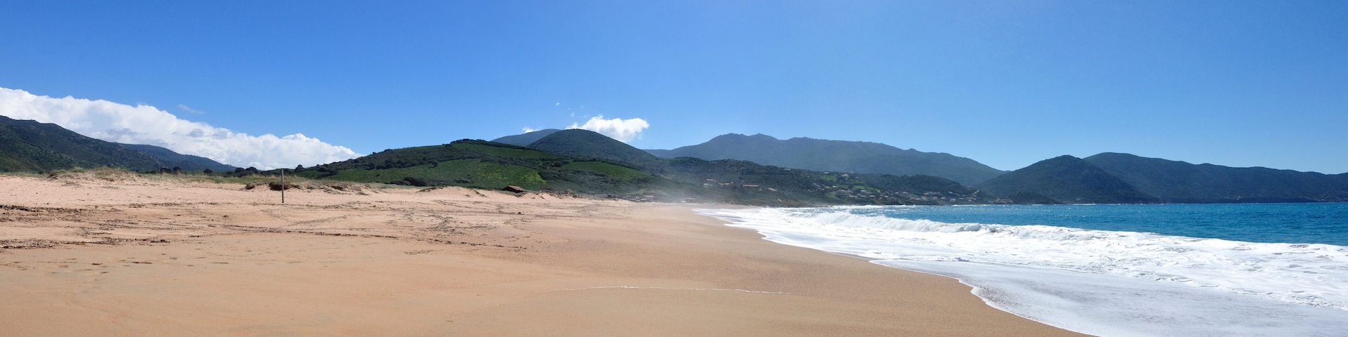 Una playa con cielo despejado, olas del océano y montañas verdes al fondo.
