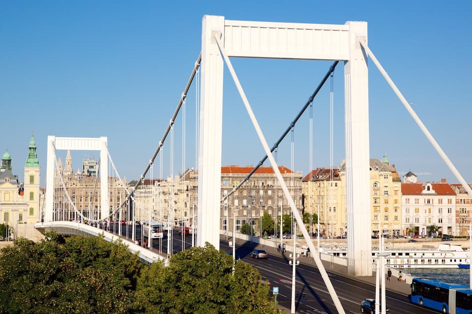 Un pont aux tours blanches et aux câbles apparents, une rivière et des bâtiments historiques aux flèches vertes en arrière-plan.