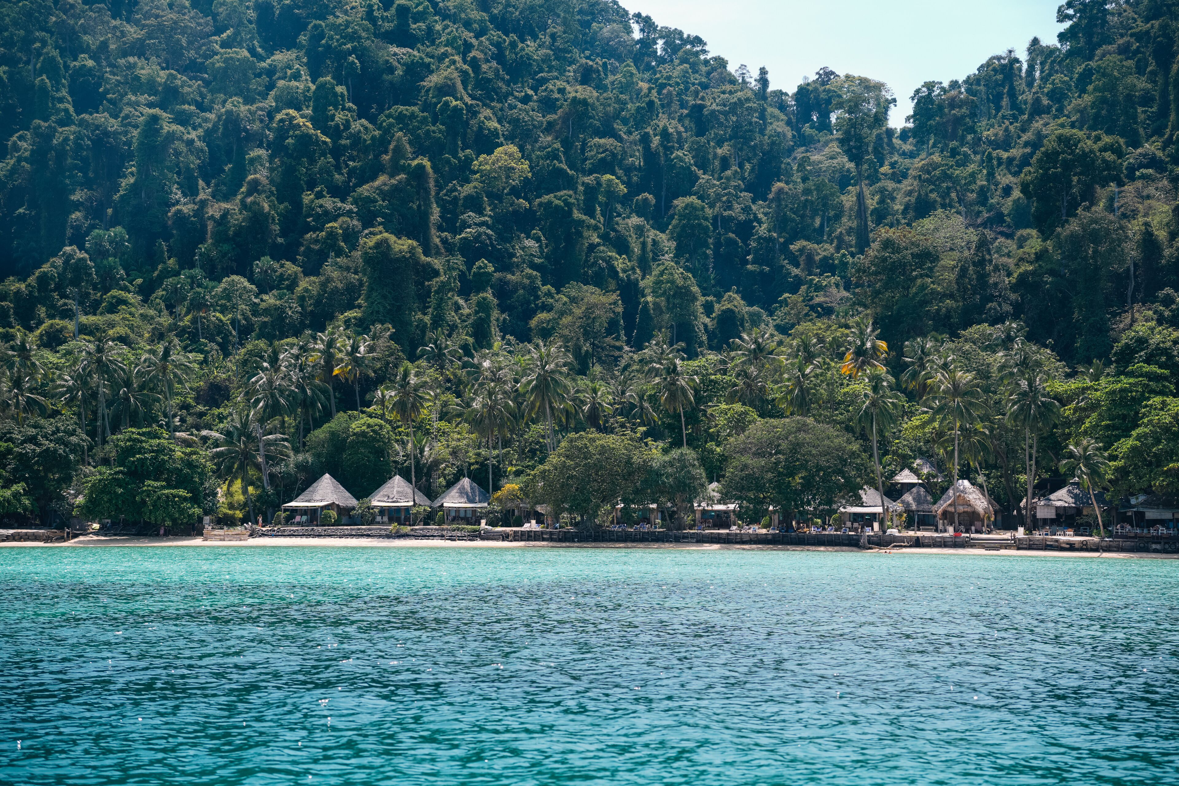 A tropical beach with thatched-roof huts and dense forested hills in the background.
