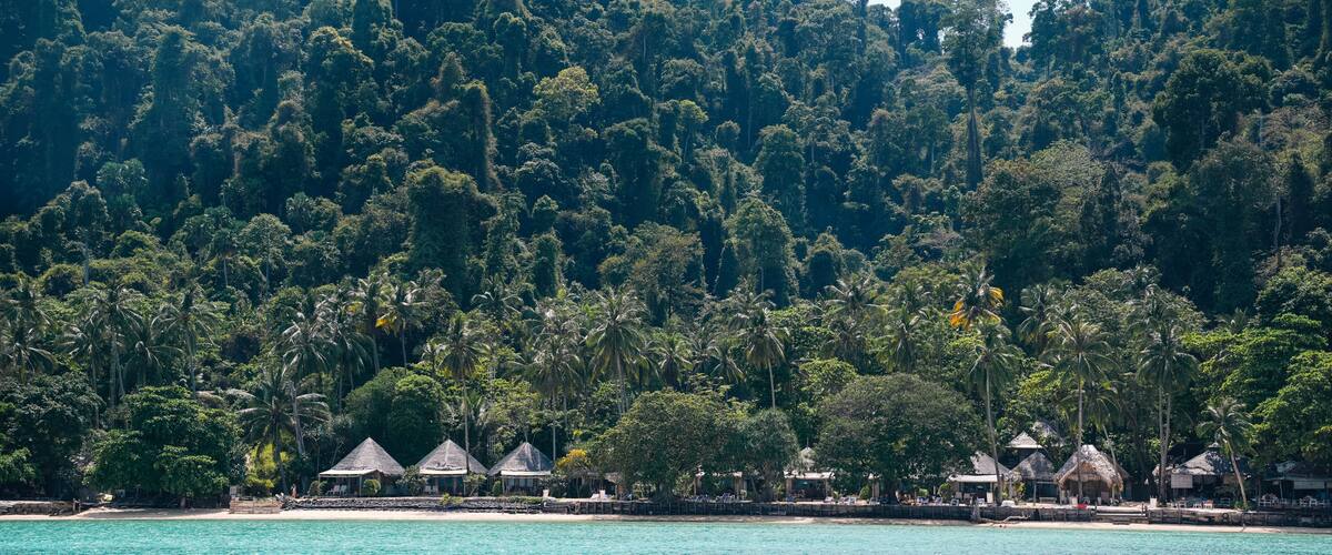 A tropical beach with thatched-roof huts and dense forested hills in the background.