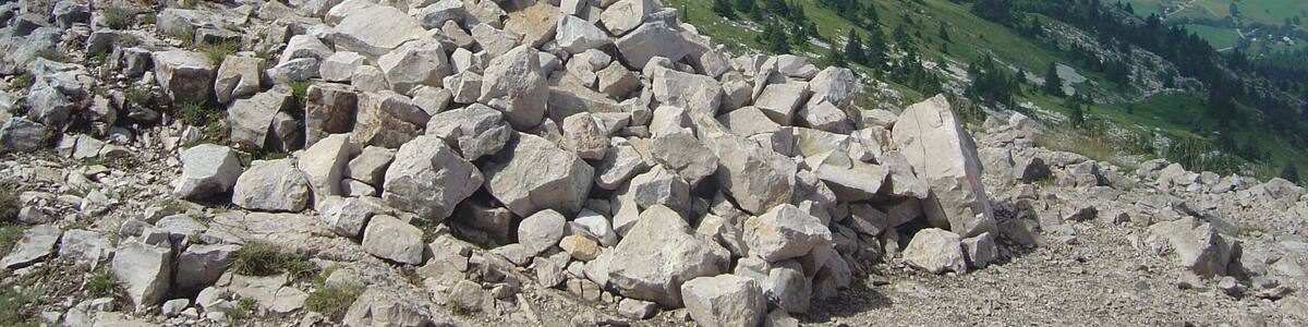 In the foreground on a hilltop there is a cross on a pile of stone. In the background there is a village in a valley, and in the distance there are mountains