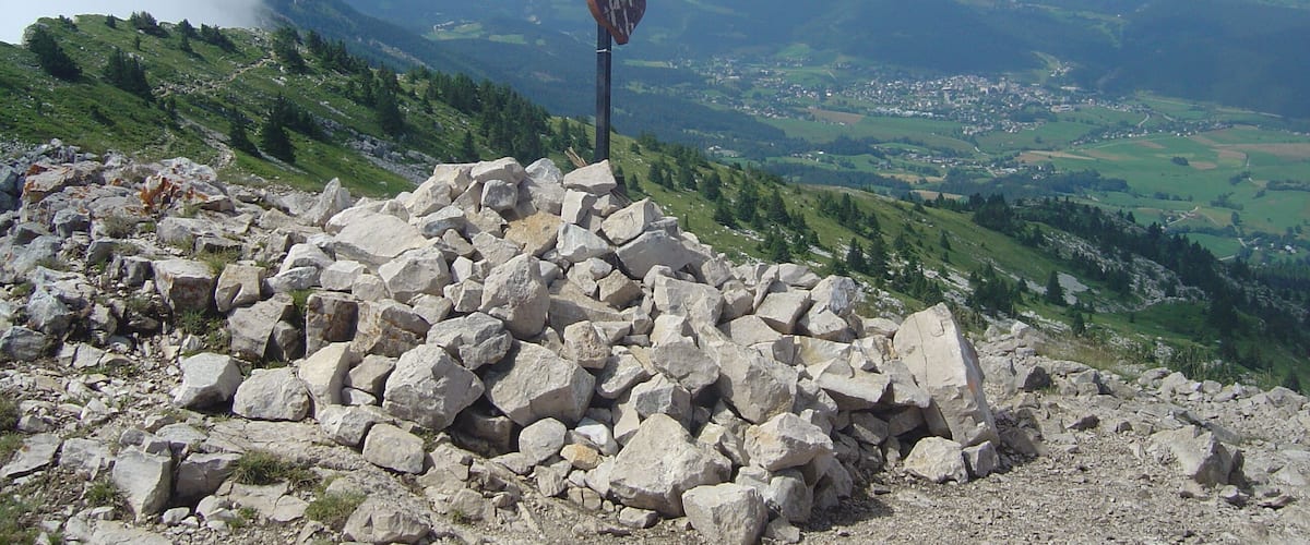 In the foreground on a hilltop there is a cross on a pile of stone. In the background there is a village in a valley, and in the distance there are mountains
