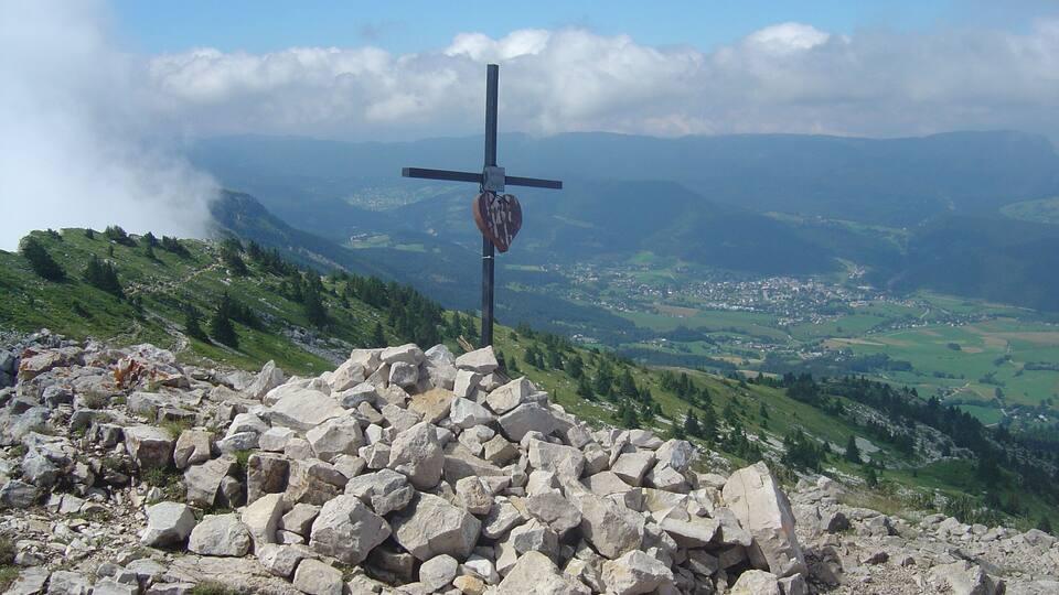A cross with a heart-shaped plaque on a rocky hilltop, overlooking a valley with scattered houses and green fields.