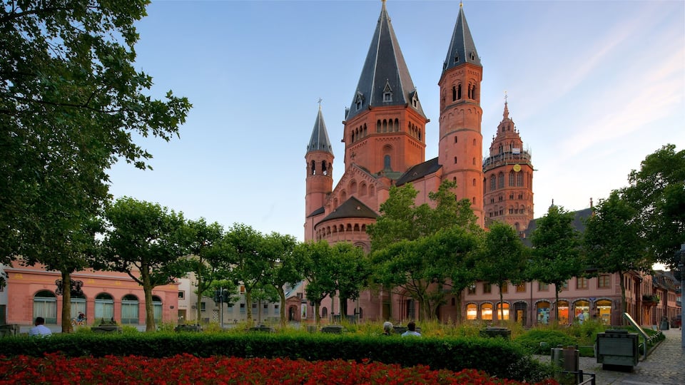 A historic building with multiple towers and a clock tower, surrounded by trees and red flowers.