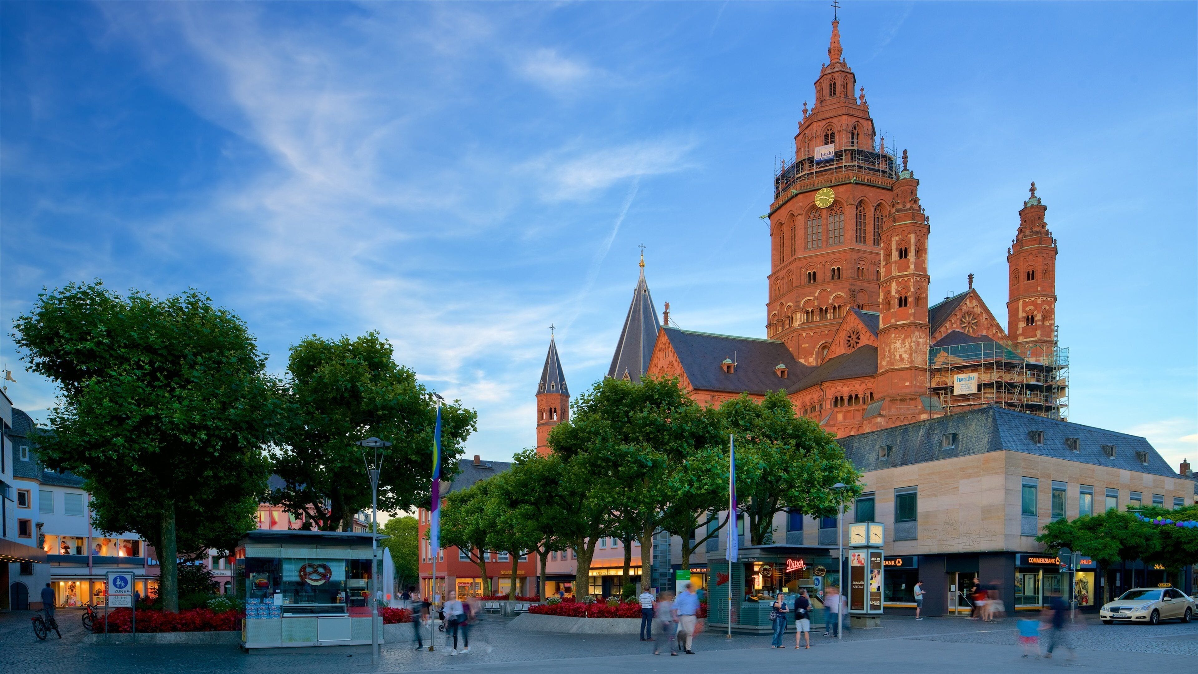 A historic church with a clock tower, surrounded by trees and a bustling square with people and shops.