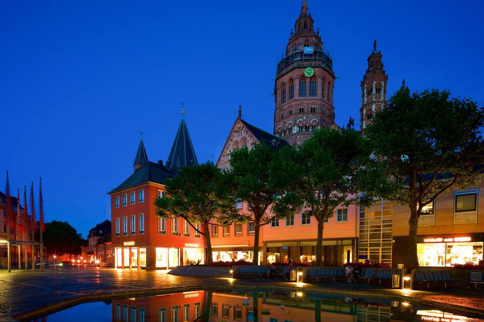 A historic town square at dusk with a prominent church tower, reflecting buildings, and a reflective water feature.