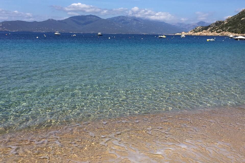 A clear coastal beach with boats in the water and mountains in the background.