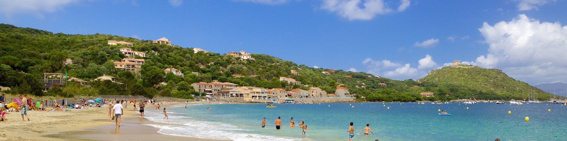 A beach with people swimming and walking along the shore, surrounded by buildings and hills.