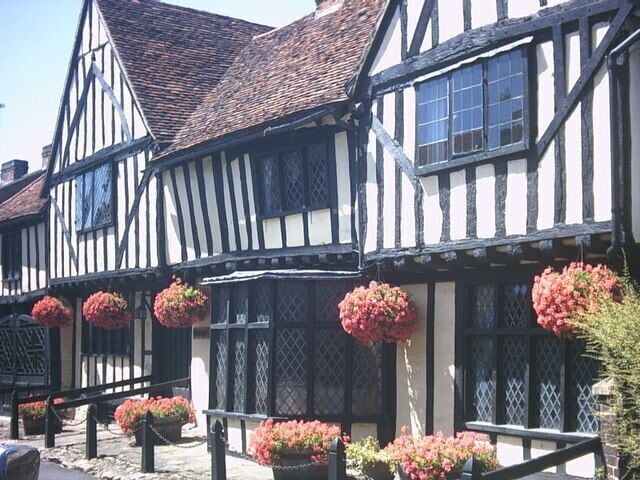 A Tudor-style building with black and white timber framing, red flowers in hanging baskets, and a cobblestone street.