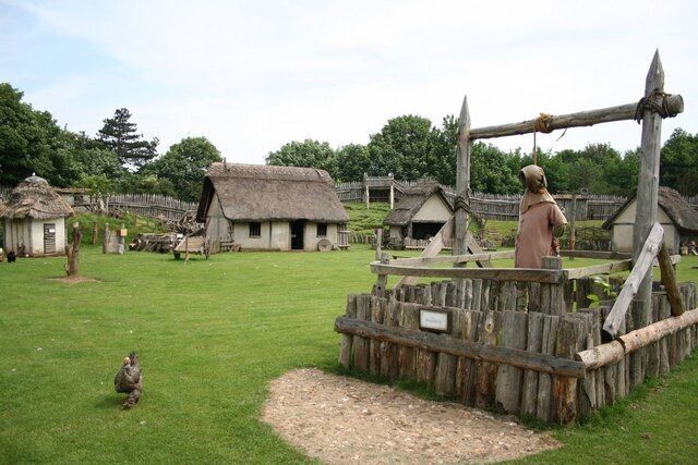 A historical village with thatched-roof houses, a wooden mill, and a fenced area with a sign.