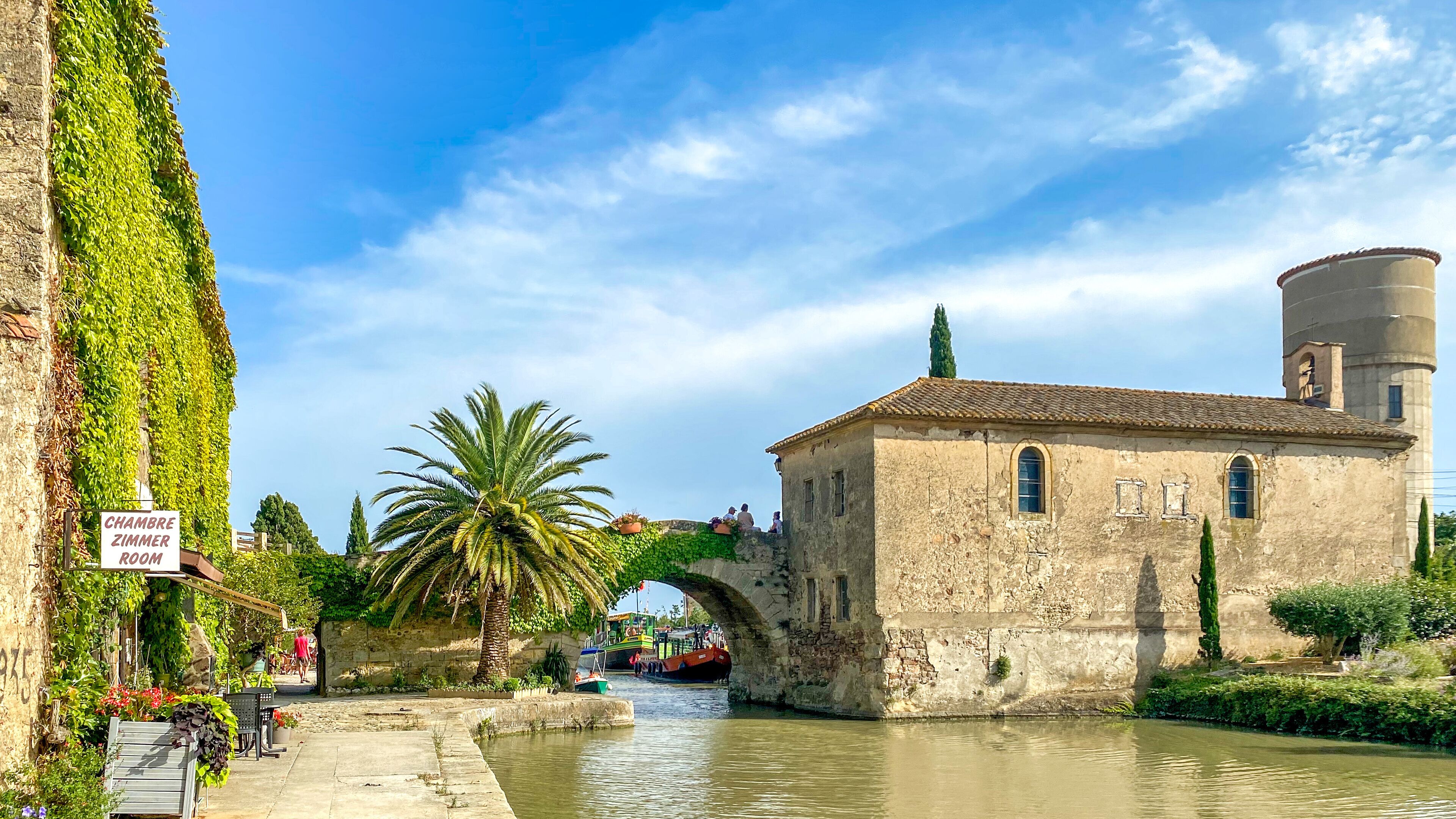 A historic building with a sign reading "Chambre Zimmer Room," a palm tree, a stone bridge, and a canal with boats.