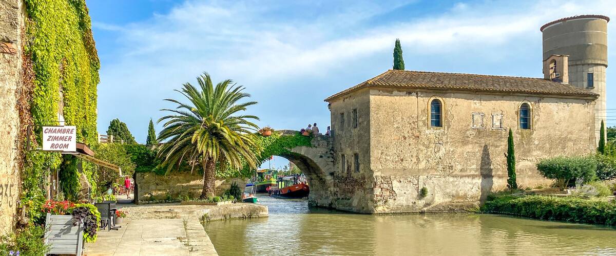A historic building with a sign reading "Chambre Zimmer Room," a palm tree, a stone bridge, and a canal with boats.