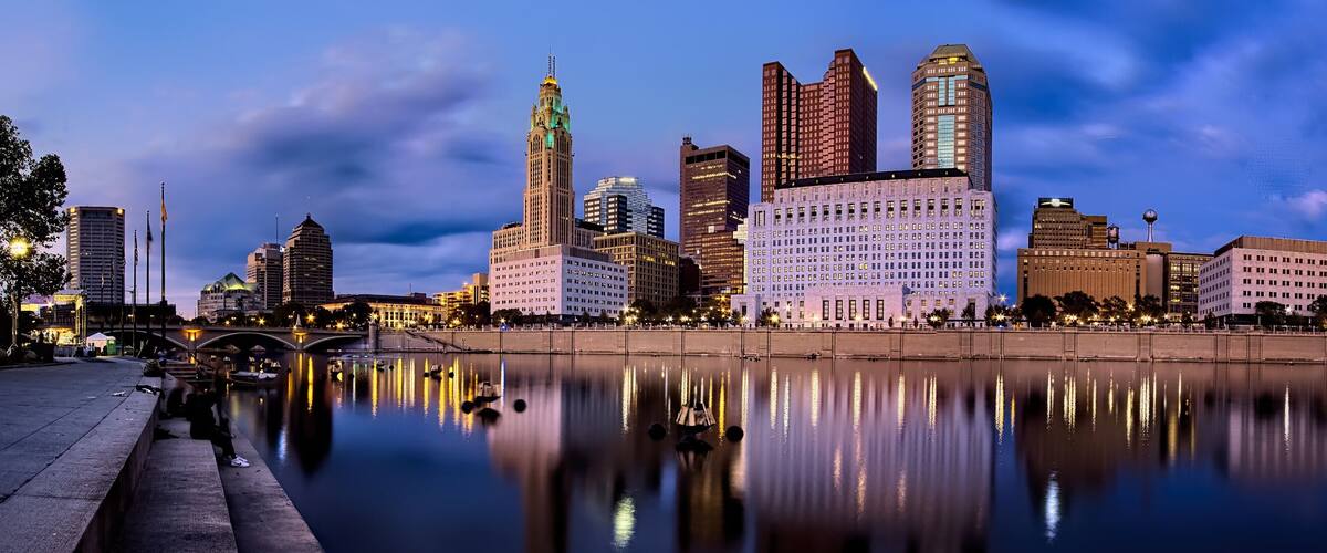 A city skyline at dusk with illuminated buildings reflected in a calm river.