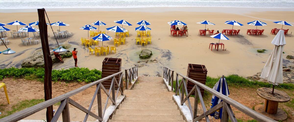 A beach with a wooden staircase leading down to the sand, surrounded by blue and white umbrellas, yellow chairs, and red tables.