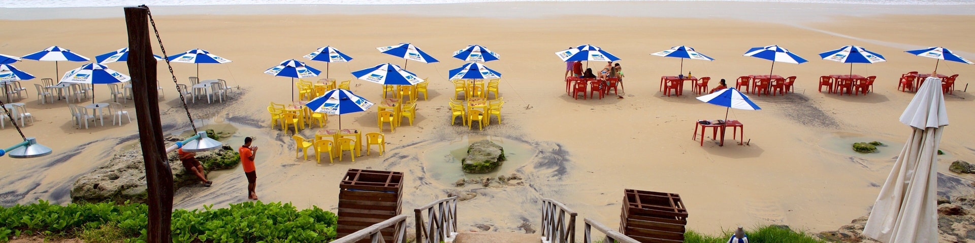 A beach with a wooden staircase leading down to the sand, surrounded by blue and white umbrellas, yellow chairs, and red tables.