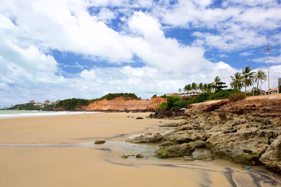 A sandy beach with rocky outcrops, palm trees, and buildings in the background.