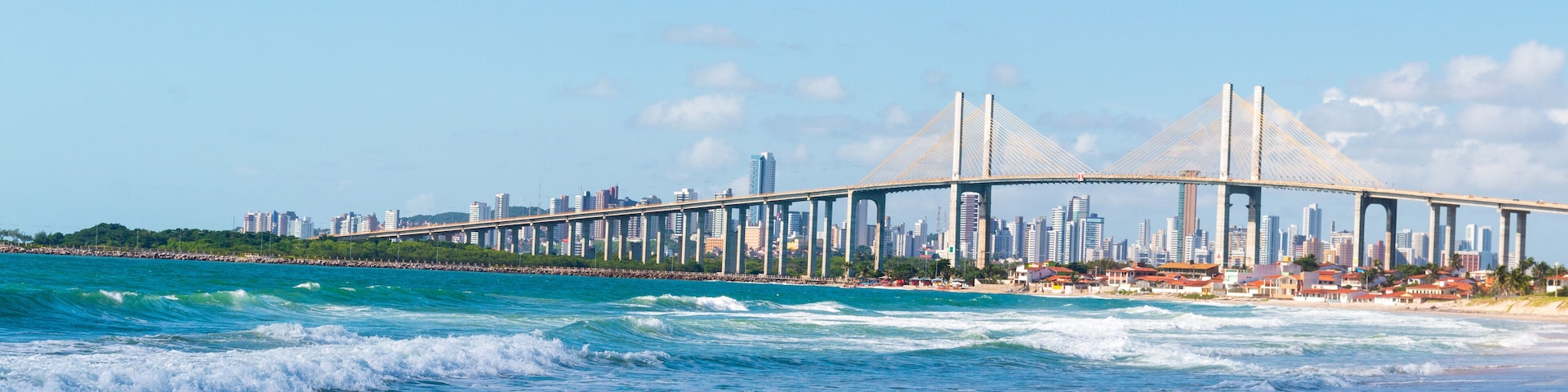 Paisagem costeira com uma ponte imponente sobre as águas, uma praia de areia e o horizonte da cidade ao fundo.