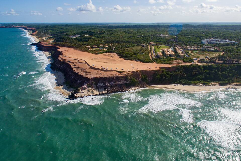 Vista aérea de uma área costeira com praia, falésias e uma floresta densa.