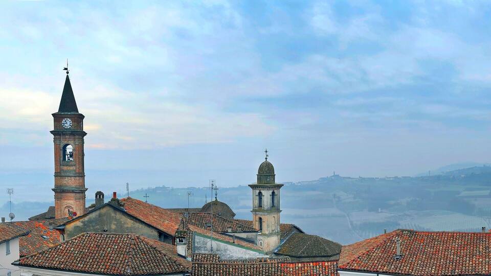 A town with red-tiled roofs, a prominent clock tower, and a distant view of rolling hills.