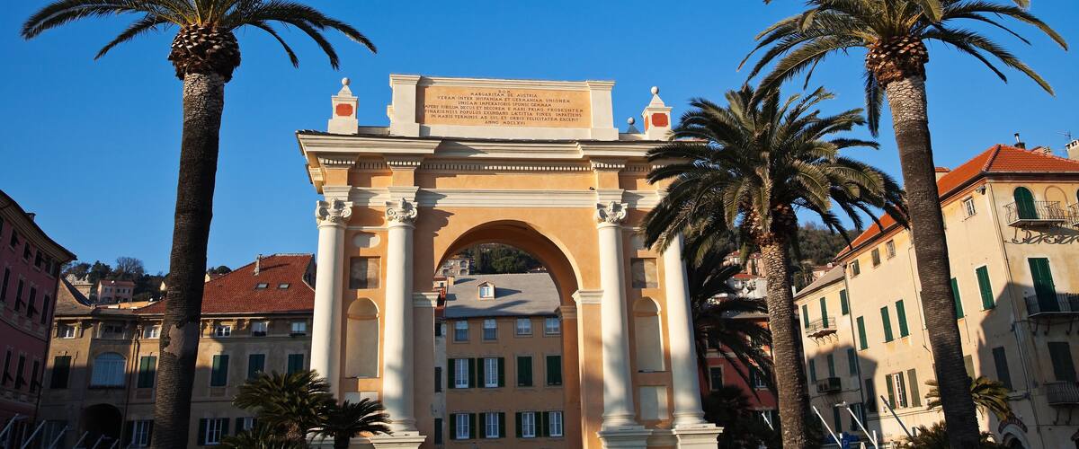 A historic archway with inscriptions, flanked by palm trees and buildings with red-tiled roofs.