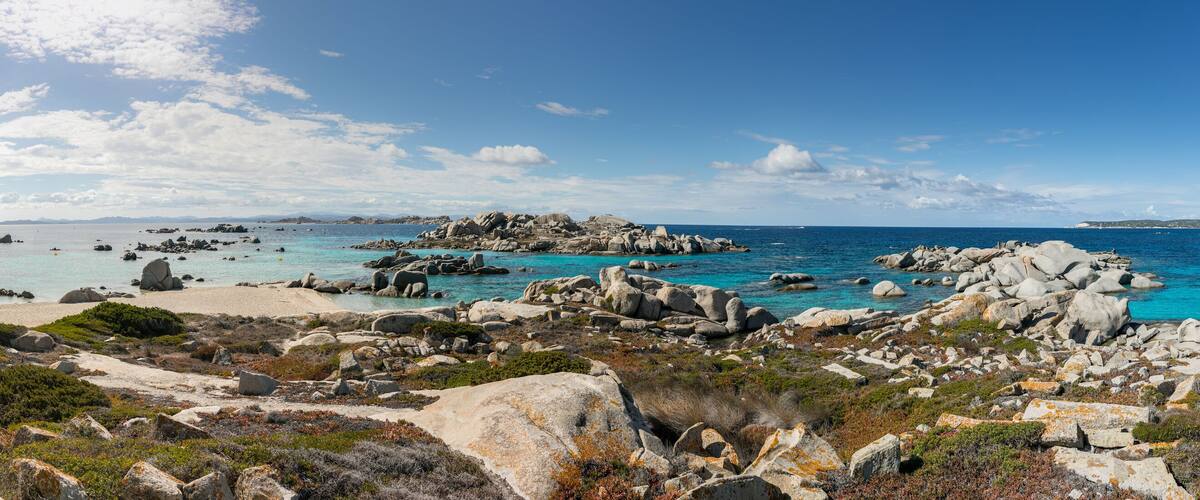 A rocky coastline with clear blue waters, a distant bridge, and a hilly landscape.