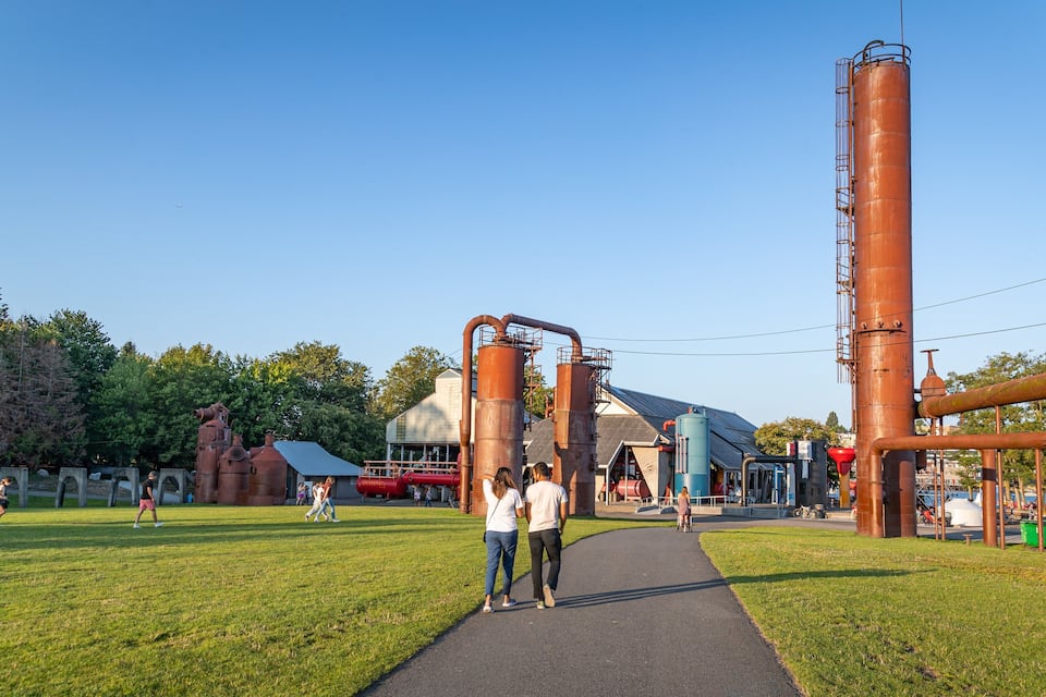 A park with a paved path, large industrial structures, and a building with a sloped roof.