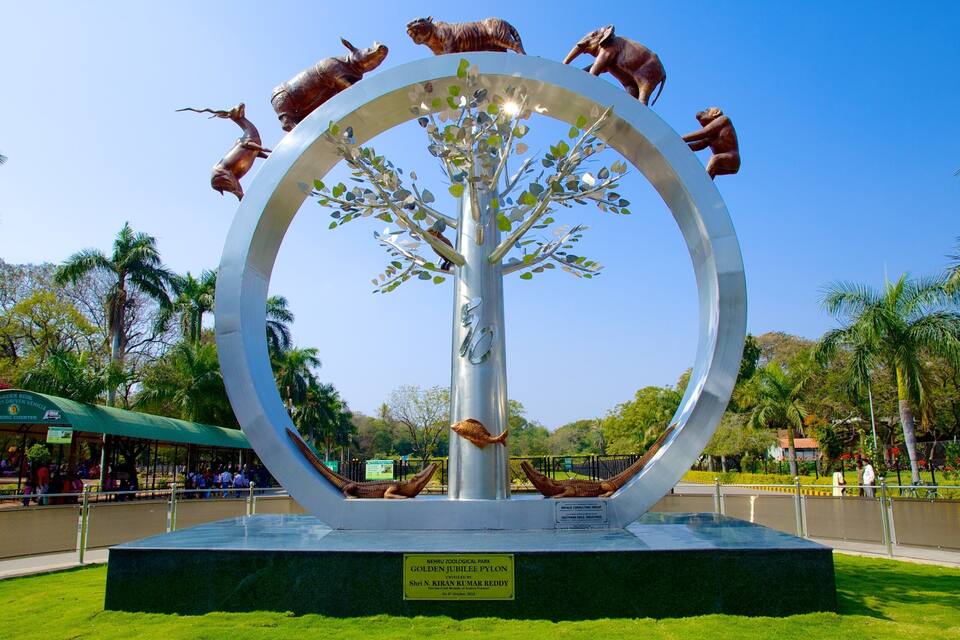Uma escultura circular com uma árvore no centro, cercada por figuras de animais, localizada em um parque com palmeiras e um edifício com telhado verde ao fundo.