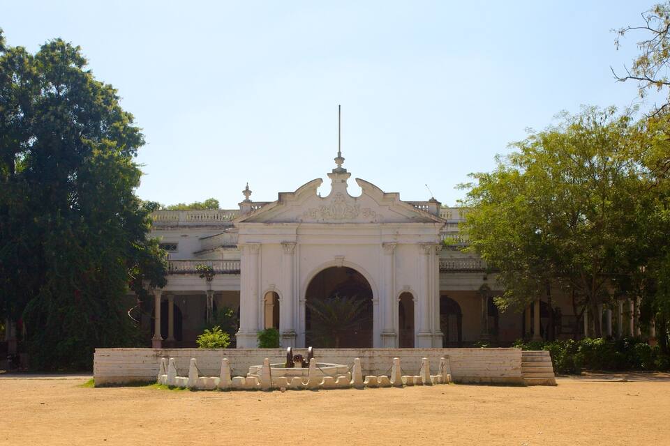 A colonial-style building with a central archway, surrounded by a sandy courtyard and trees.