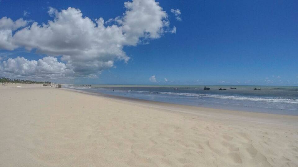 A wide sandy beach with gentle waves, a clear blue sky, and a few boats in the water.