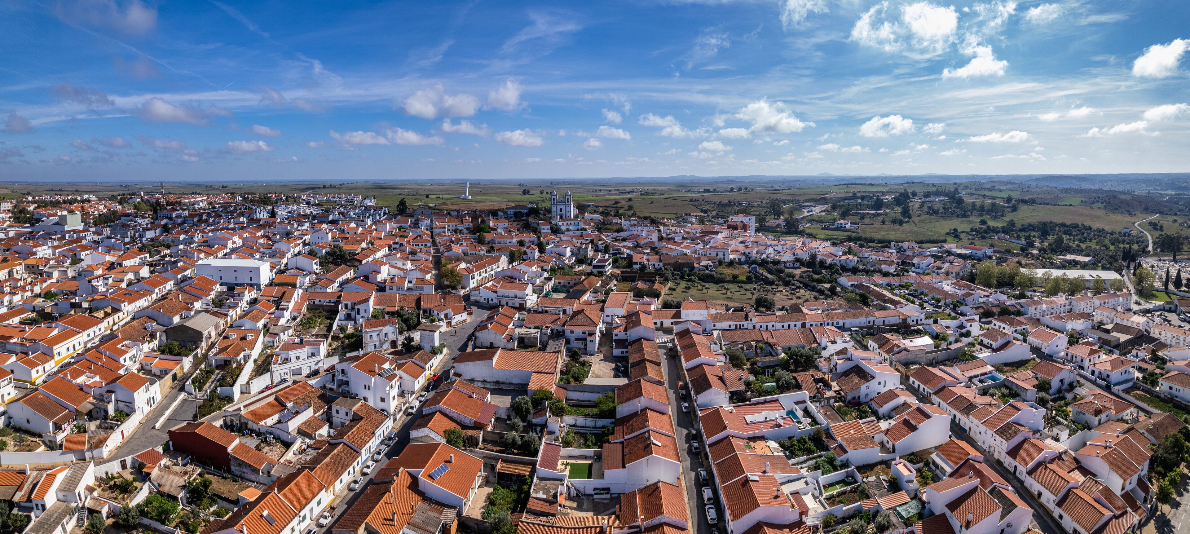 A coastal town with white buildings and red roofs, surrounded by green fields and a lighthouse in the distance.