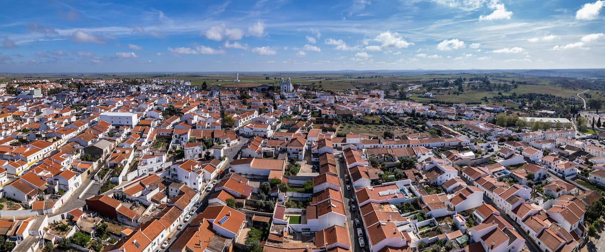 A coastal town with white buildings and red roofs, surrounded by green fields and a lighthouse in the distance.
