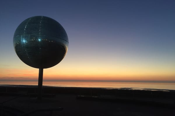 Uma grande escultura esférica e espelhada em um píer, com o céu ao pôr do sol ao fundo.