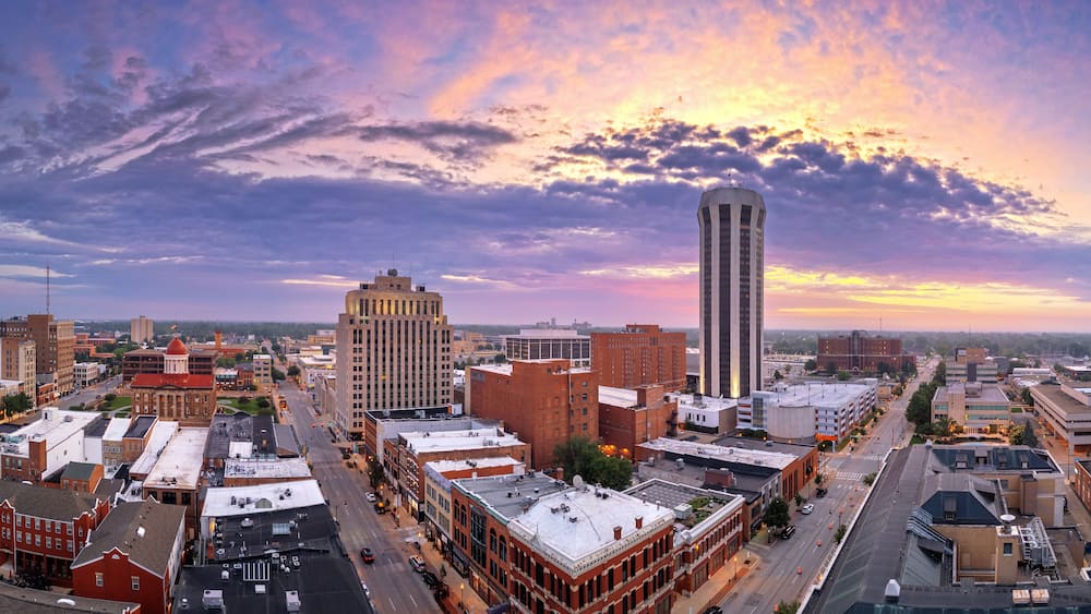 A cityscape with a prominent tall tower, a historic building with a dome, and a variety of other buildings under a colorful sunset sky.