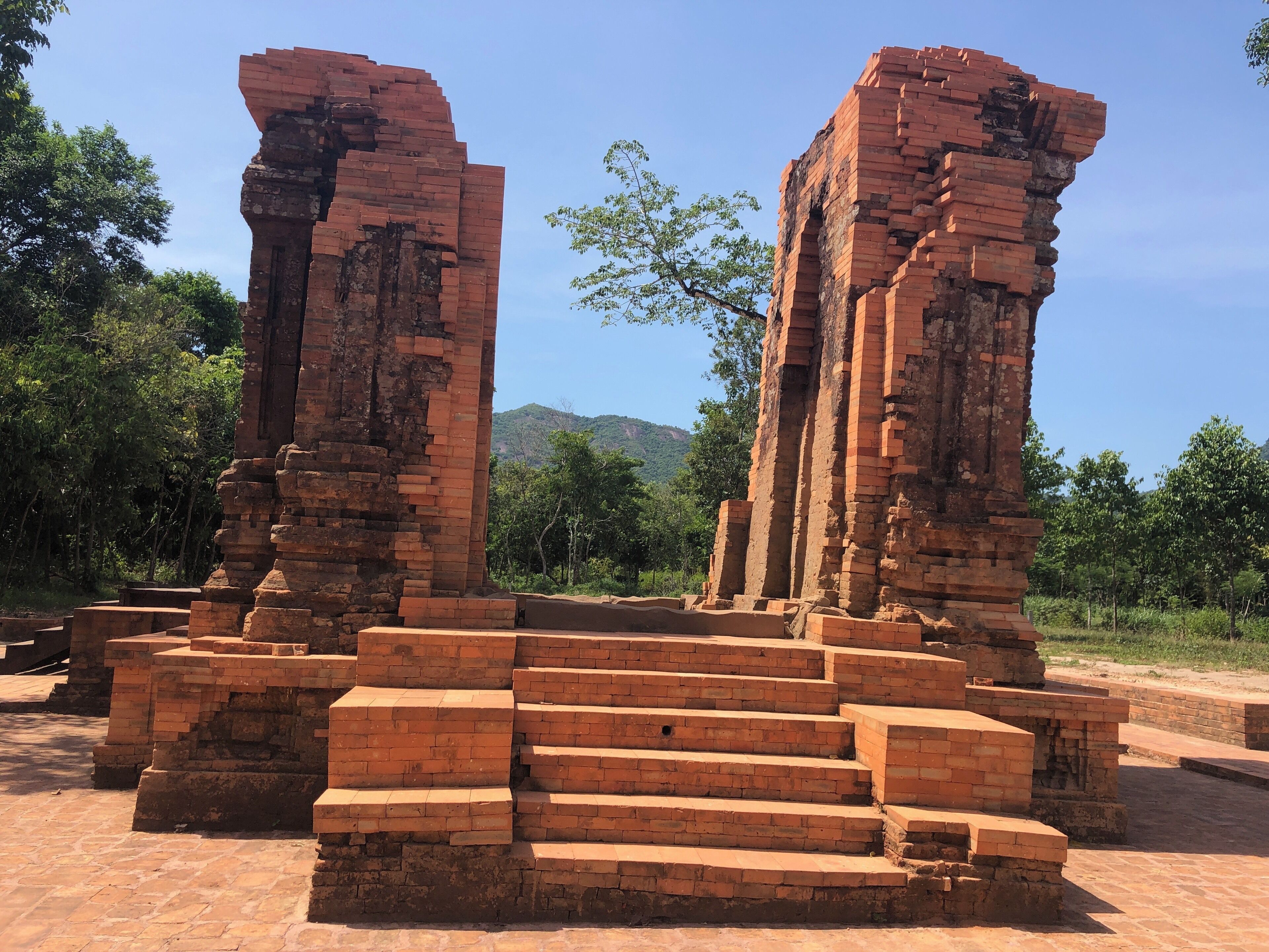 Ancient brick ruins with steps leading up to them, surrounded by trees and hills in the background.