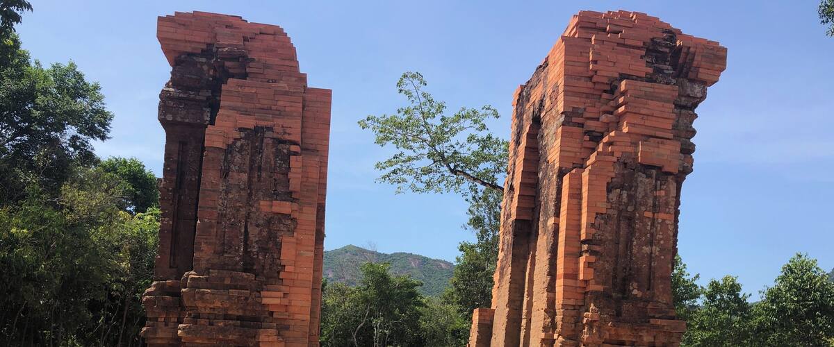 Ancient brick ruins with steps leading up to them, surrounded by trees and hills in the background.