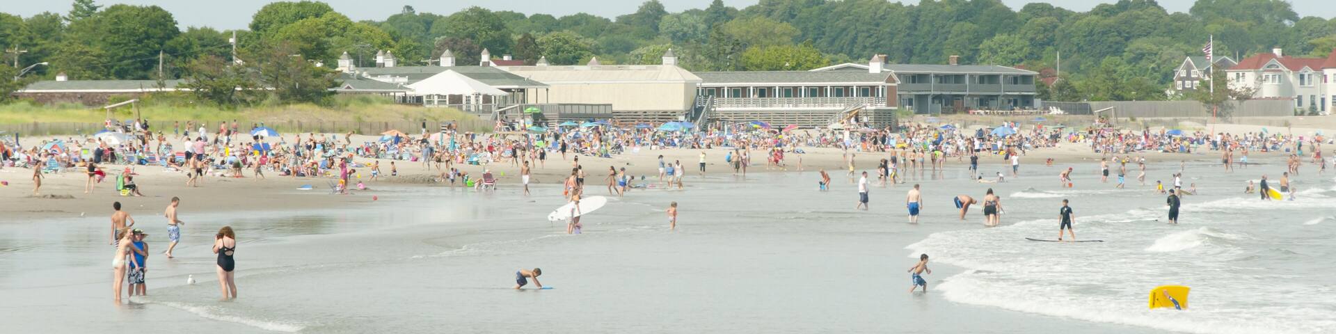 A crowded beach with people swimming and surfing, a lifeguard tower, and buildings in the background.