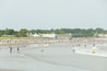 A crowded beach with people swimming and surfing, a lifeguard tower, and buildings in the background.
