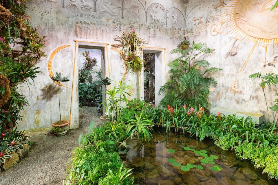 Un jardin intérieur luxuriant, avec une grande variété de plantes, un petit étang parsemé de nénuphars et un mur orné de sculptures décoratives.
