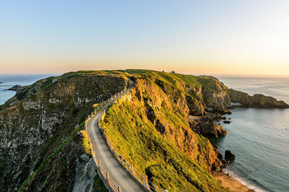 Eine Küstenstraße schlängelt sich an einer Klippe entlang mit Blick auf das Meer und Booten in der Ferne.
